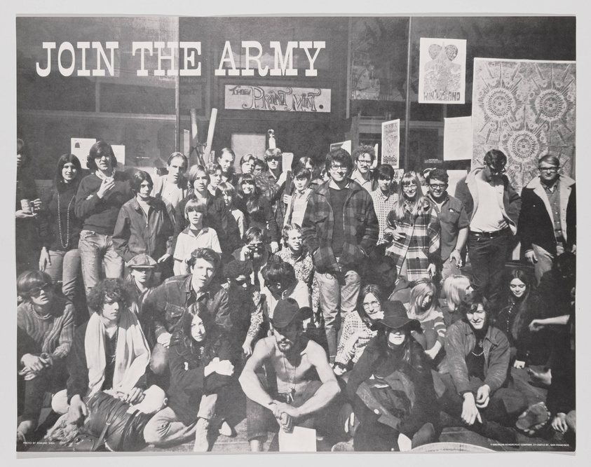 A black and white photograph of a large group of young people, some seated and others standing, in front of a building with a sign that reads "JOIN THE ARMY." The individuals display a variety of fashion styles indicative of the late 1960s or early 1970s, with several posters visible in the background. The atmosphere suggests a protest or gathering, with some participants holding signs, and the overall mood appears informal and possibly countercultural.