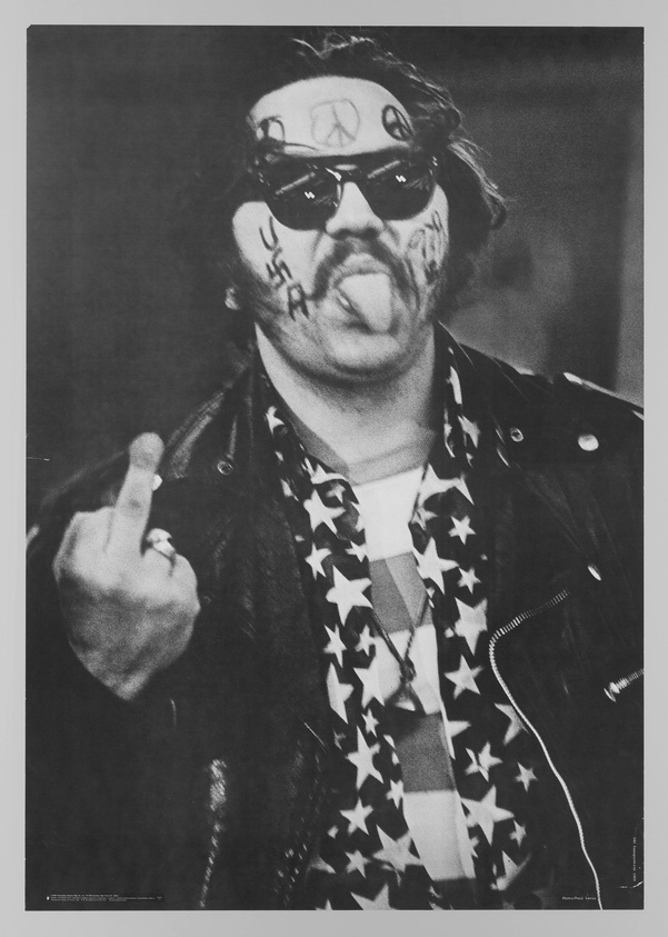 Black and white photograph of a man sticking out his tongue and making an obscene gesture with his hand. He wears sunglasses, a bandana with a peace sign, and a leather jacket over a shirt with a star-spangled pattern. His face is marked with various symbols and letters.