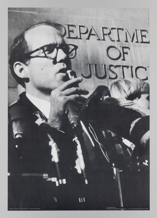 A white man wearing glasses has a microphone touching his lips and stands in front of a building titled "Department of Justice"