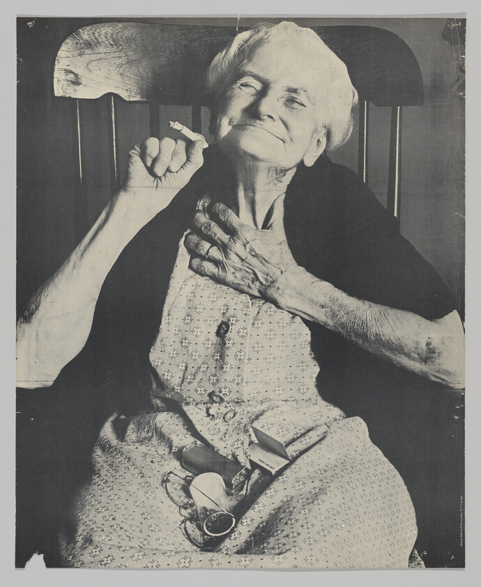 Elderly woman sits smiling with a cigarette in one hand and glasses on her lap.