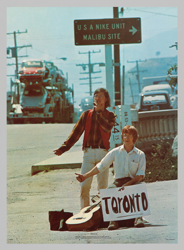 Two young men hitchhiking at the roadside, one holding a guitar and a sign reading "Toronto."