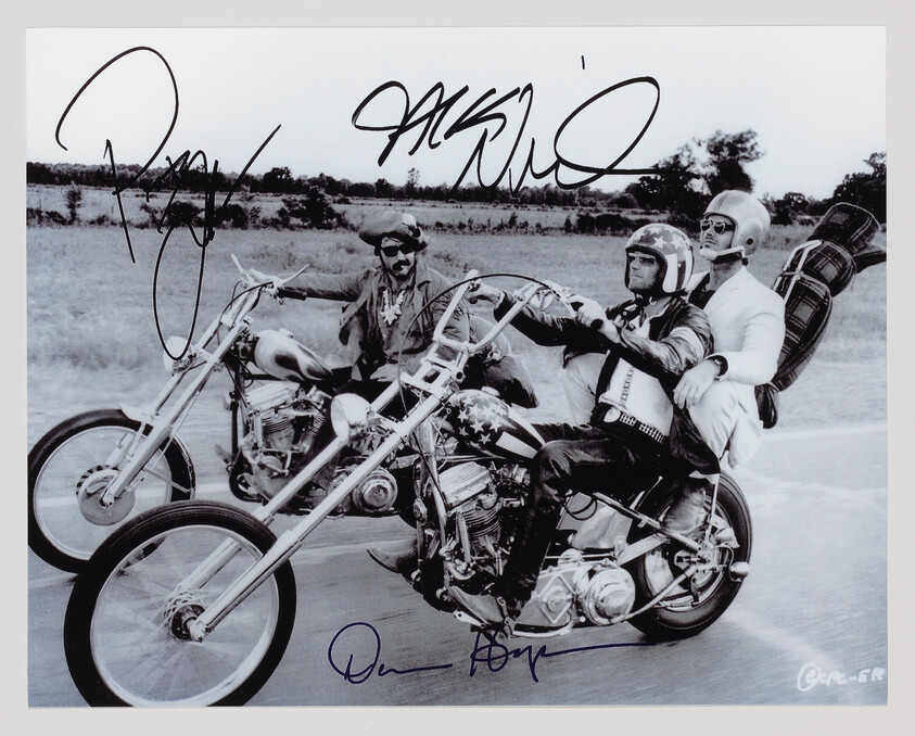 Three motorcyclists ride chopper-style bikes on a road, one passenger wearing white carrying a bag.