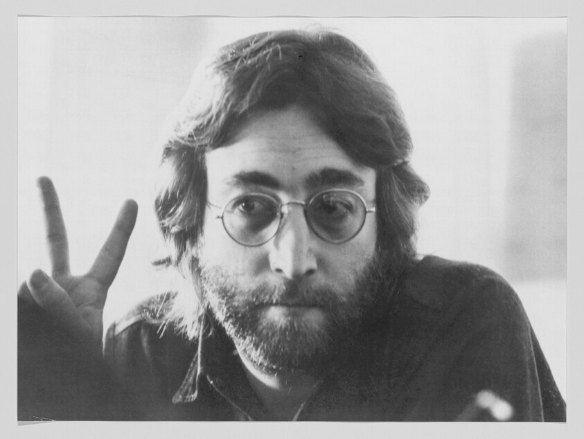 Bearded man wearing round glasses holds up two fingers in a peace sign.