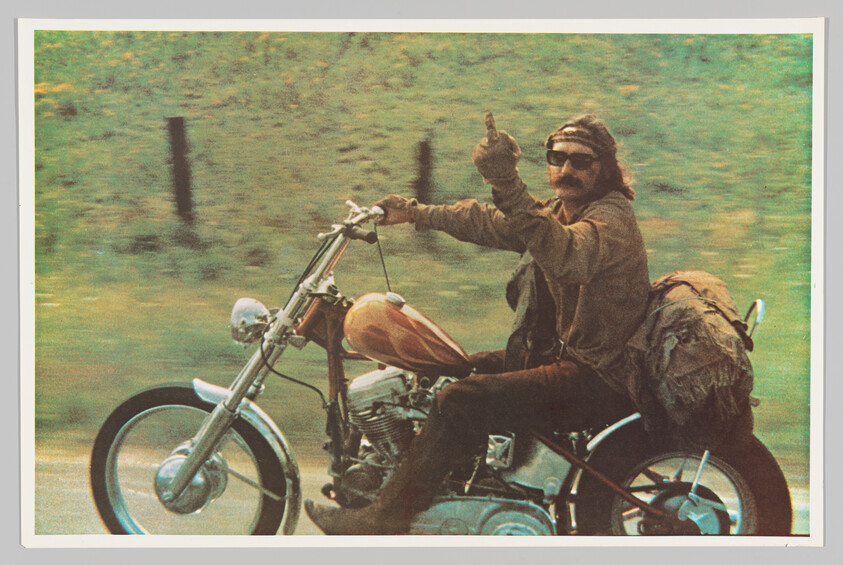 Man on a motorcycle rides past a country road while giving a thumbs-up.