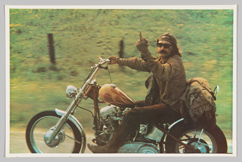 A man riding a classic chopper-style motorcycle gives a peace sign with his right hand. He is wearing a leather jacket, sunglasses, and a headband, and has a rugged appearance. The motorcycle is equipped with a round headlight and high handlebars, and there's a large bag attached to the side. The background is a blur of greenery, suggesting motion.