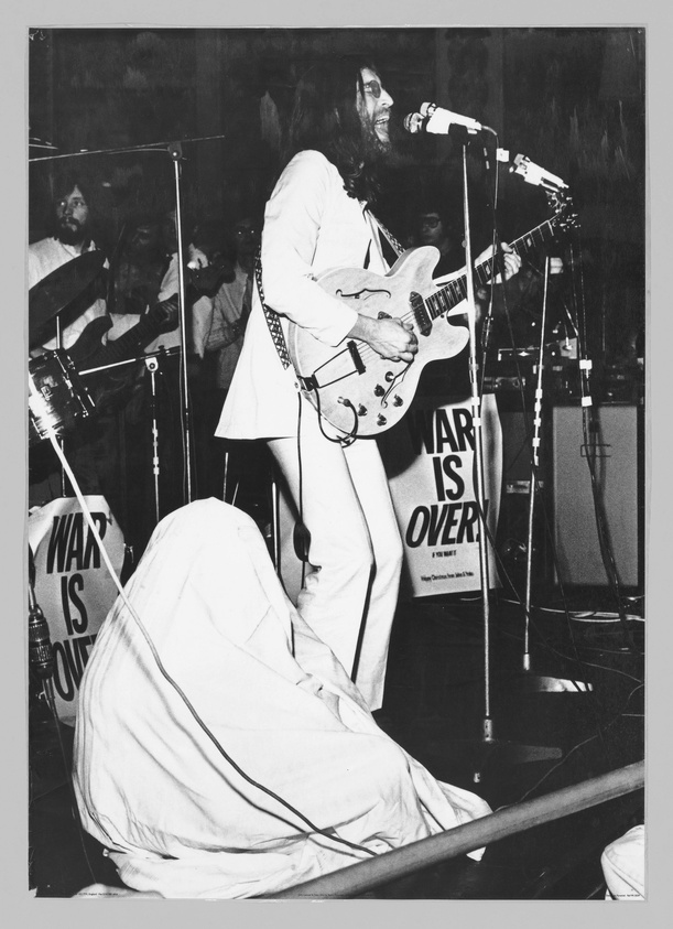 A black and white photograph of a musician with long hair and a beard, playing an electric guitar and singing into a microphone on a stage. The musician is wearing a white suit, and there is a sign with the message "WAR IS OVER! IF YOU WANT IT" in the background. Another person is partially visible, covered with a white cloth, sitting on the stage floor. There are microphone stands and cables around, and the audience is visible in the background.