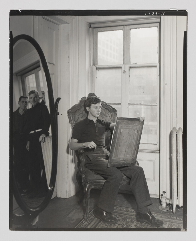 A young man sits in a chair holding a drawing board, with two people reflected in a large mirror.