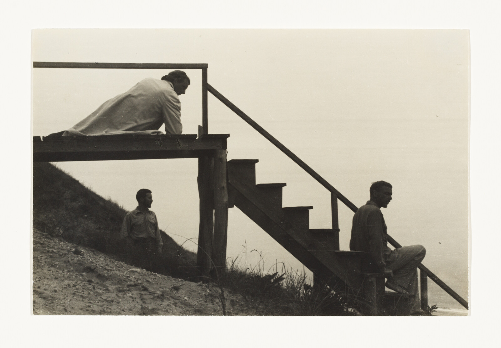 Three people on a wooden staircase by a hillside, one sitting, one standing, one leaning.