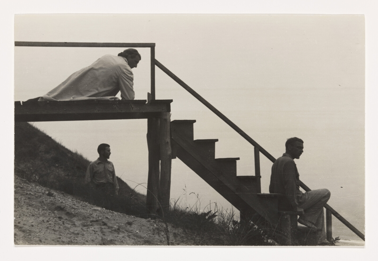 Three people on a wooden staircase by a hillside, one sitting, one standing, one leaning.