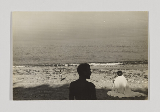 Two people sit on a beach, one wrapped in a blanket, facing the ocean.