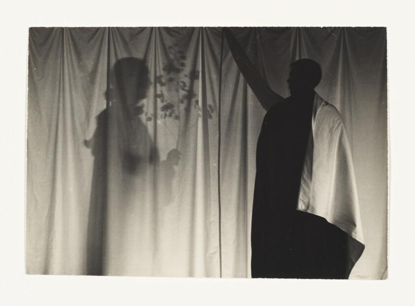 A person in a cloak reaches towards a curtain, casting a shadow of another figure.