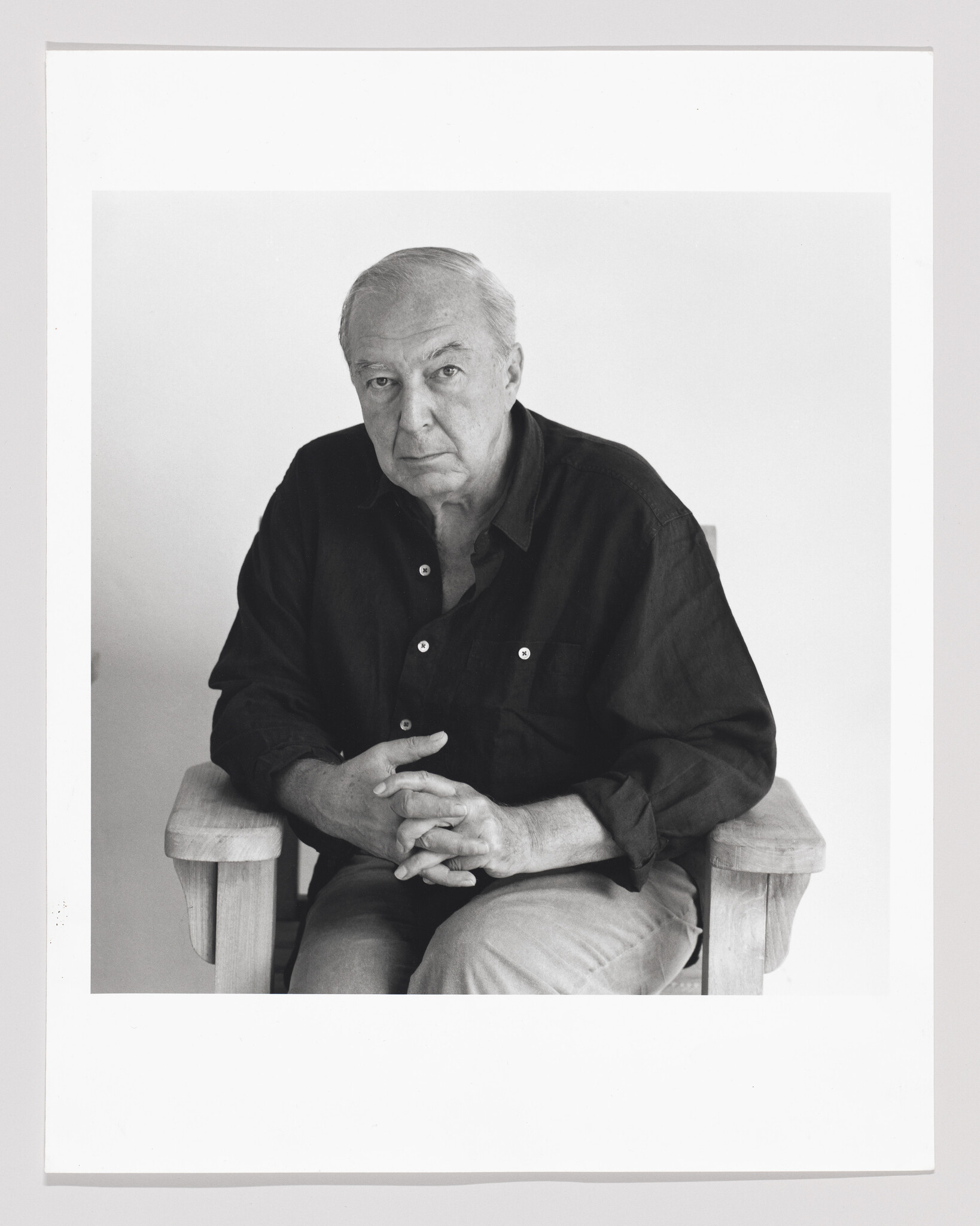 Black and white portrait of an elderly man seated on a wooden chair, looking directly at the camera with a neutral expression. He is wearing a dark shirt and light-colored pants, with his hands clasped together in his lap. The photo has a white border framing it.