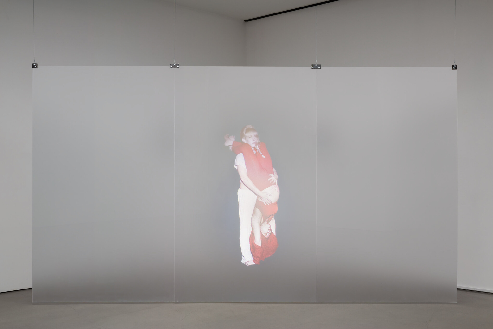 Two performers in red are entwined, one held upside down, projected onto a translucent screen.