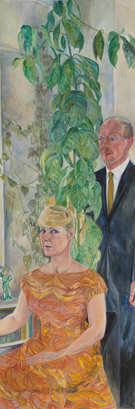 Seated woman in an orange patterned dress with a standing man in a suit behind her, tall plant between.
