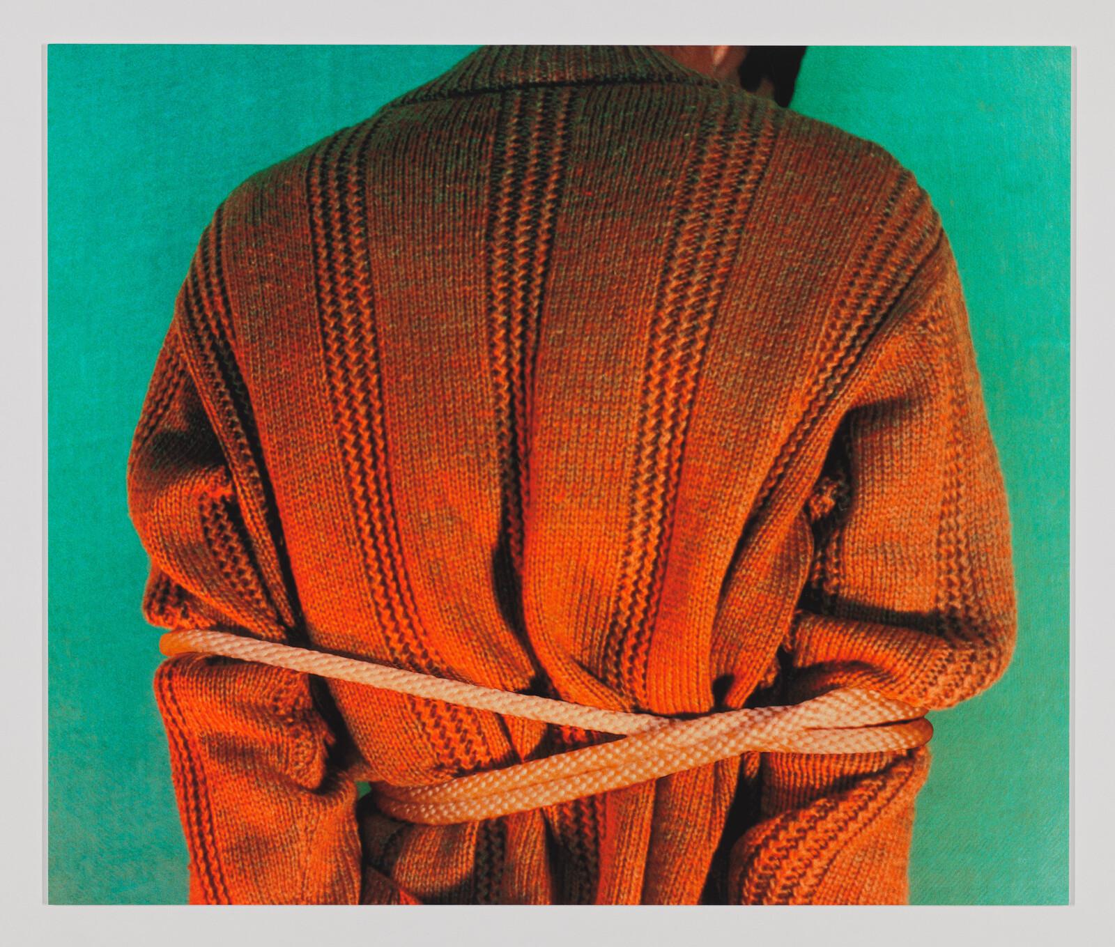 A person in an orange knit sweater stands with their arms tied behind them.