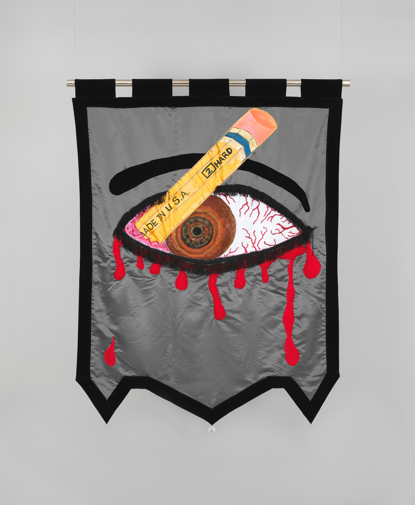 A sewn banner with a bloody brown eye stabbed by a number two pencil against a grey background. 