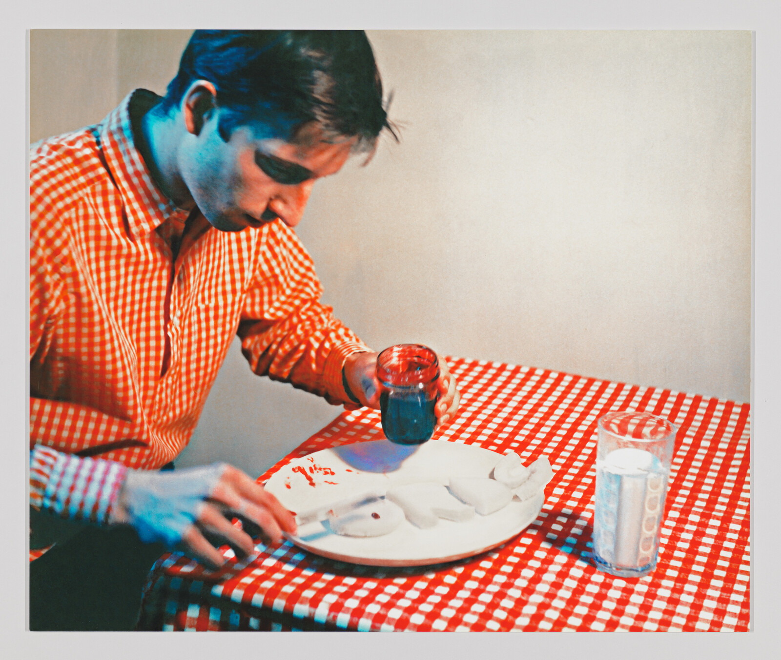 A man in a red checkered shirt paints white ceramic shapes with blue paint at a table.