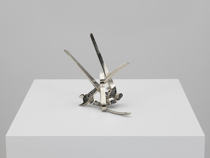 A small polished metal abstract sculpture made from overlapping kitchen utensils displayed on a white pedestal.