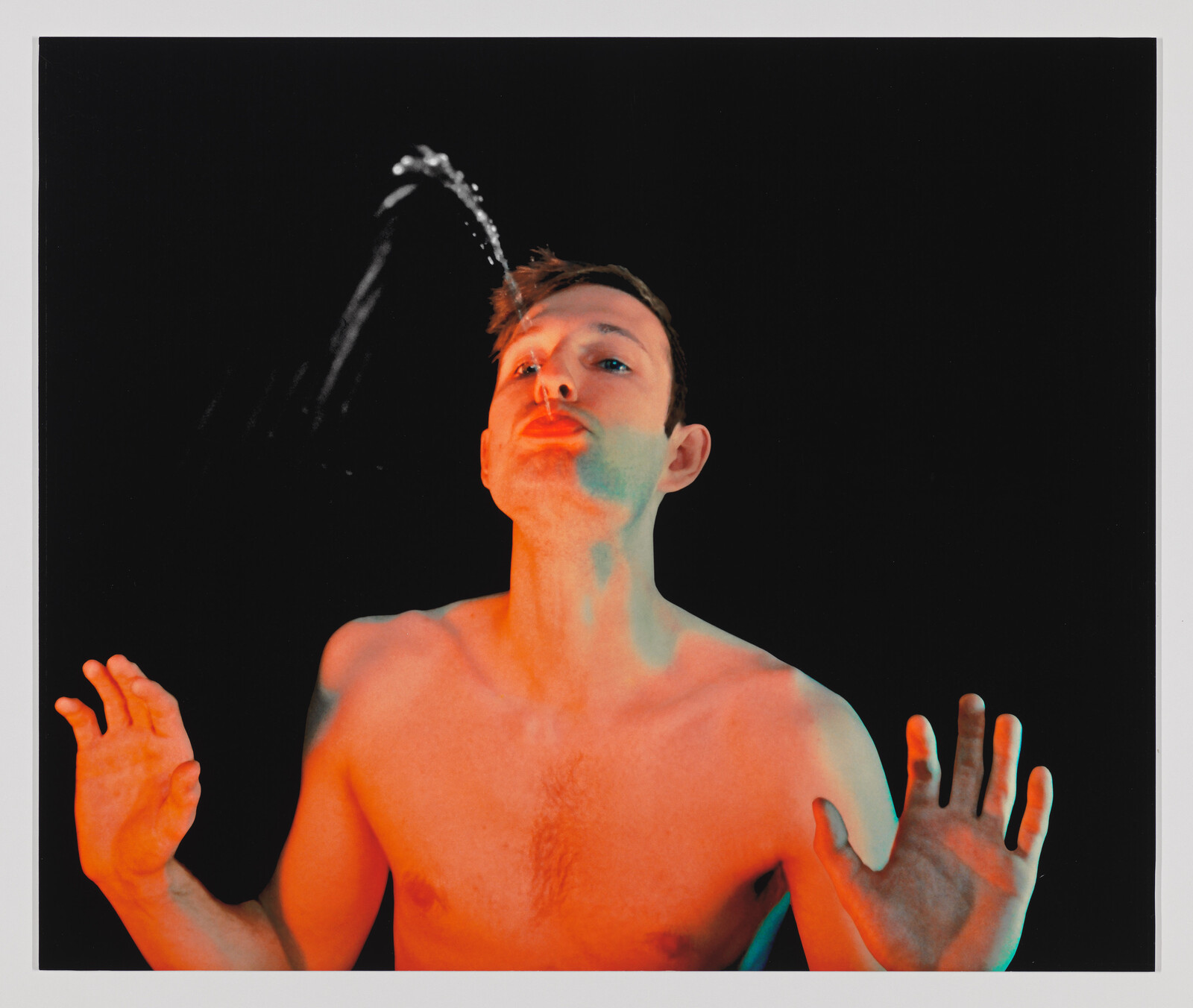 A shirtless man leans back and spits a stream of water upward against a black background.
