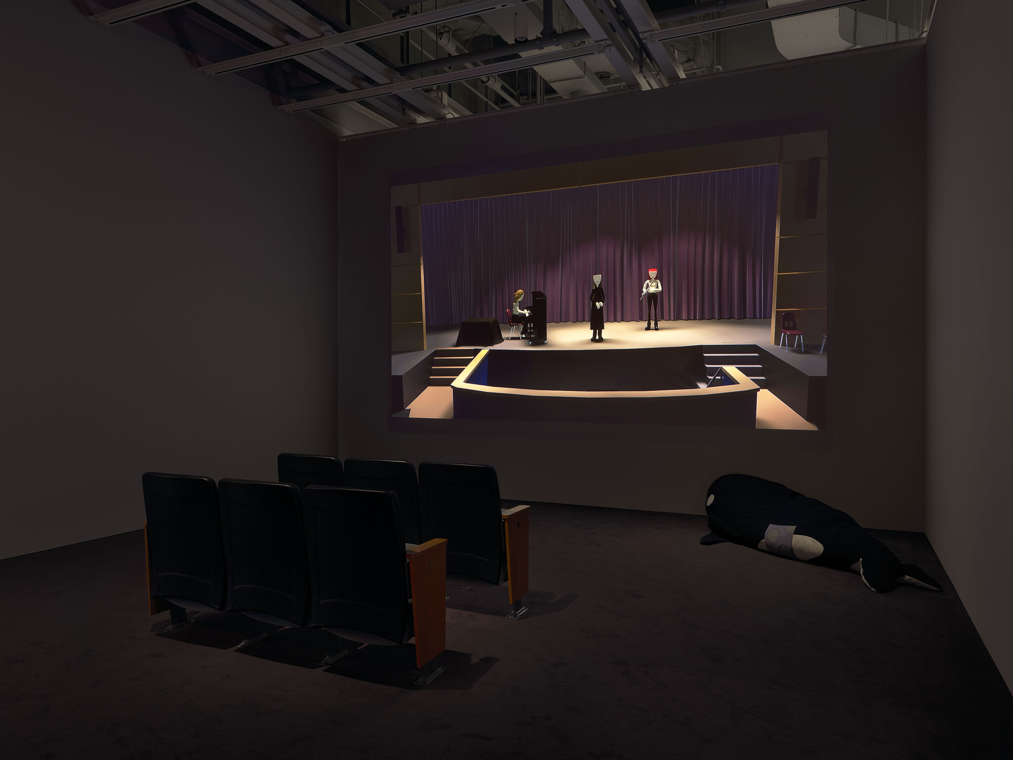 A small dark screening room showing a projected stage scene with three performers and empty seats.