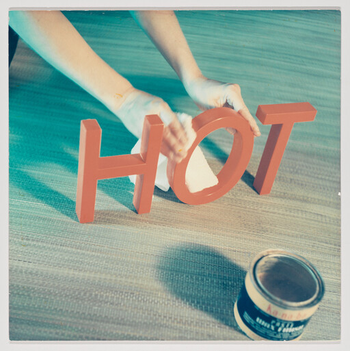 A person wipes bright red letters spelling HOT with a cloth beside an open paint can. — view 97248