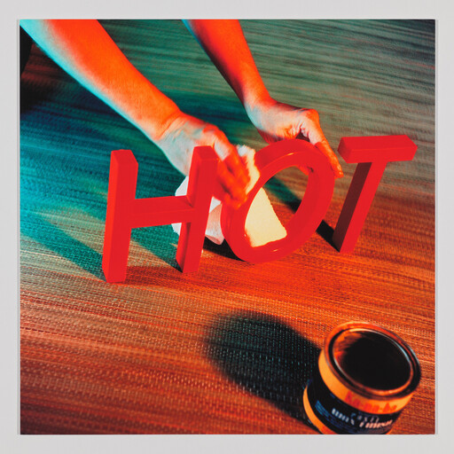 A person wipes bright red letters spelling HOT with a cloth beside an open paint can.