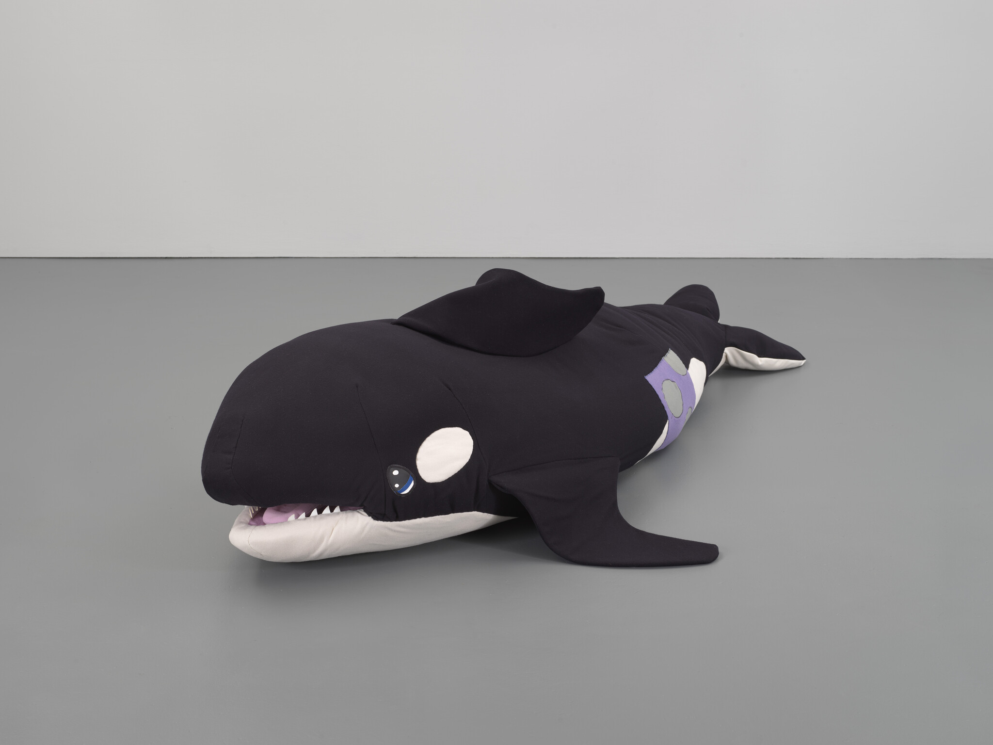 A large black and white plush orca whale lies on a gray floor with mouth slightly open.