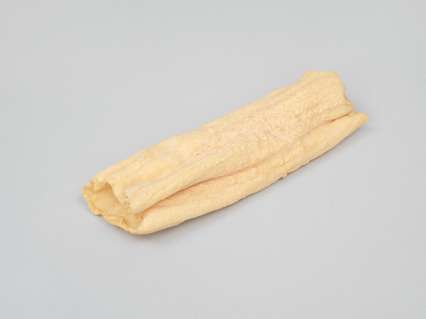 A folded strip of pale dried tofu skin (yuba) resting on a light gray background.