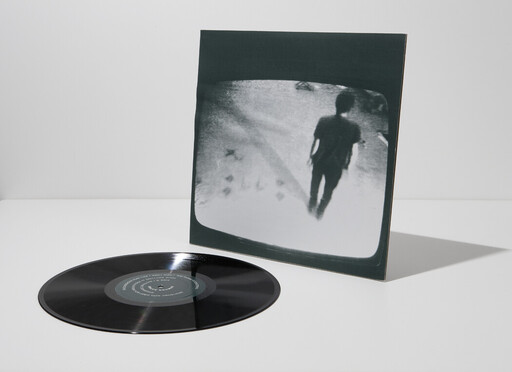 A black vinyl record lies next to its sleeve showing a grainy image of a walking person.