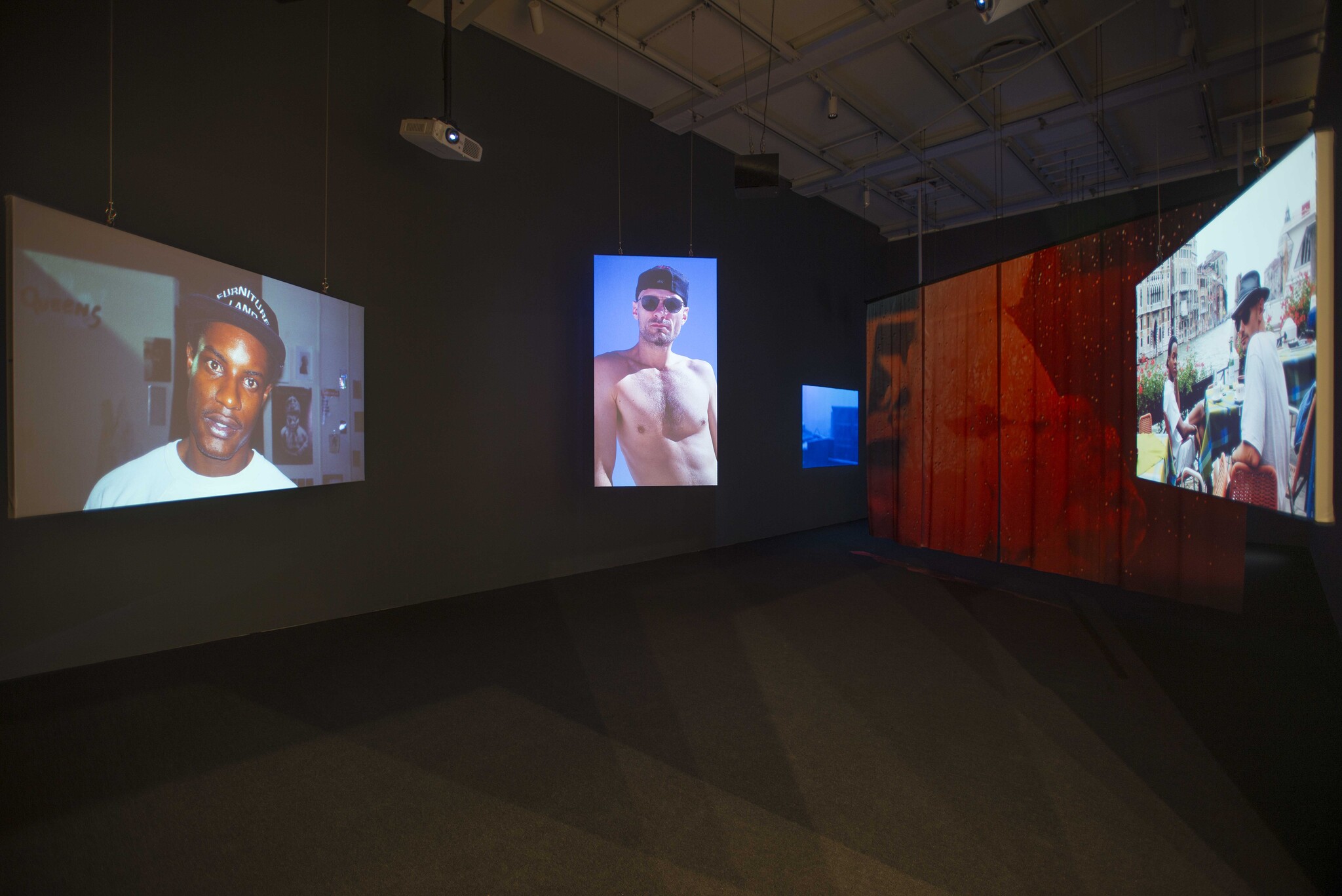 Art gallery with multiple video projections on walls, featuring portraits and scenes of people in various settings.