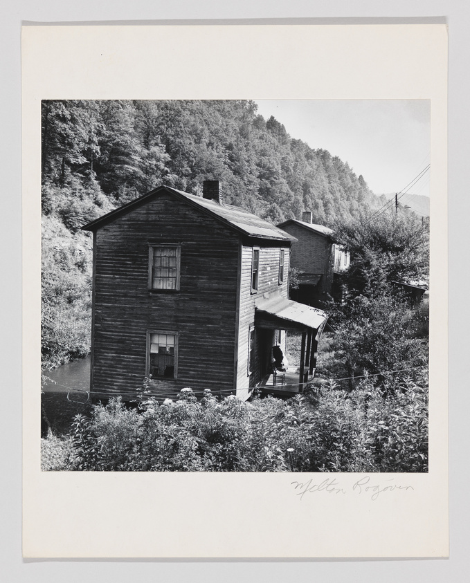 A black and white photograph depicting an old, weathered two-story wooden house with a porch. The house is situated on a slope with lush vegetation in the foreground and a dense forest in the background. There are power lines visible to the right, and another building can be seen further up the hill. The image is signed "Milton Rogovin" at the bottom right.