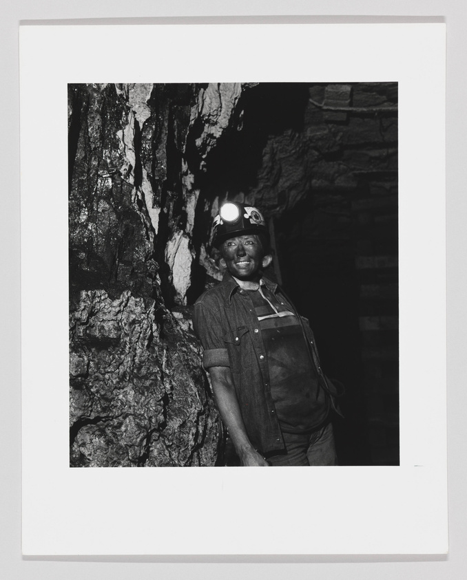 A black and white photograph of a smiling miner wearing a hard hat with a headlamp. He is standing in a rocky environment that appears to be an underground mine. The miner is dressed in work clothes with a button-up shirt and overalls.