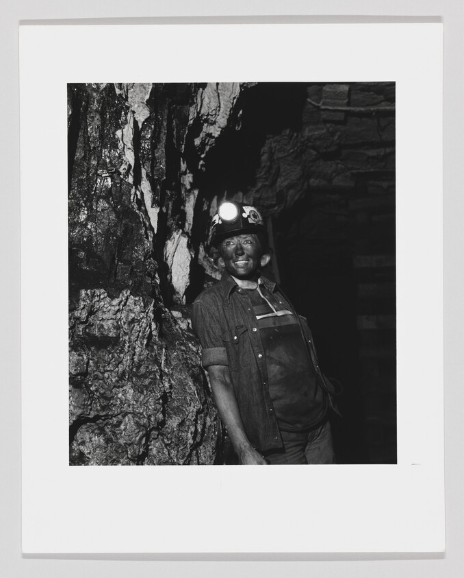 Coal miner with a lit headlamp leans against a rock wall, smiling and covered in dust.