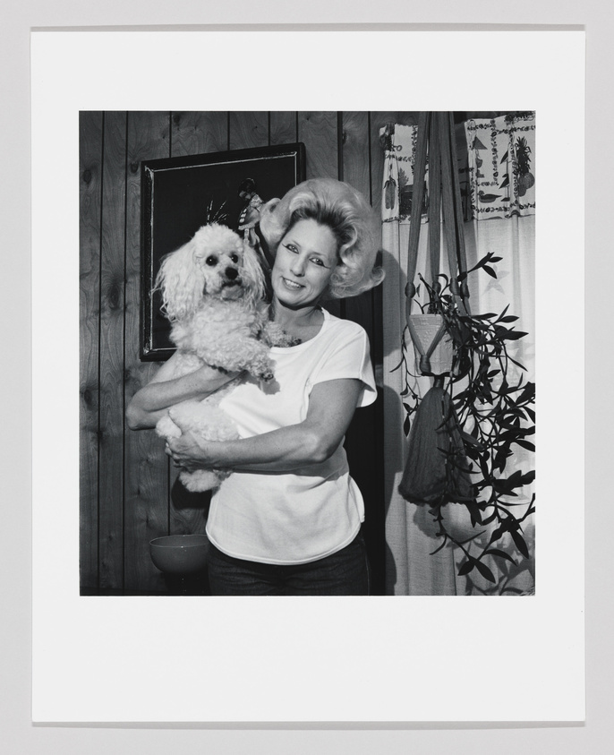 A black and white photo of a smiling woman with voluminous hair holding a fluffy dog. They are indoors, with wood-paneled walls, a framed picture, patterned curtains, and a potted plant in the background.