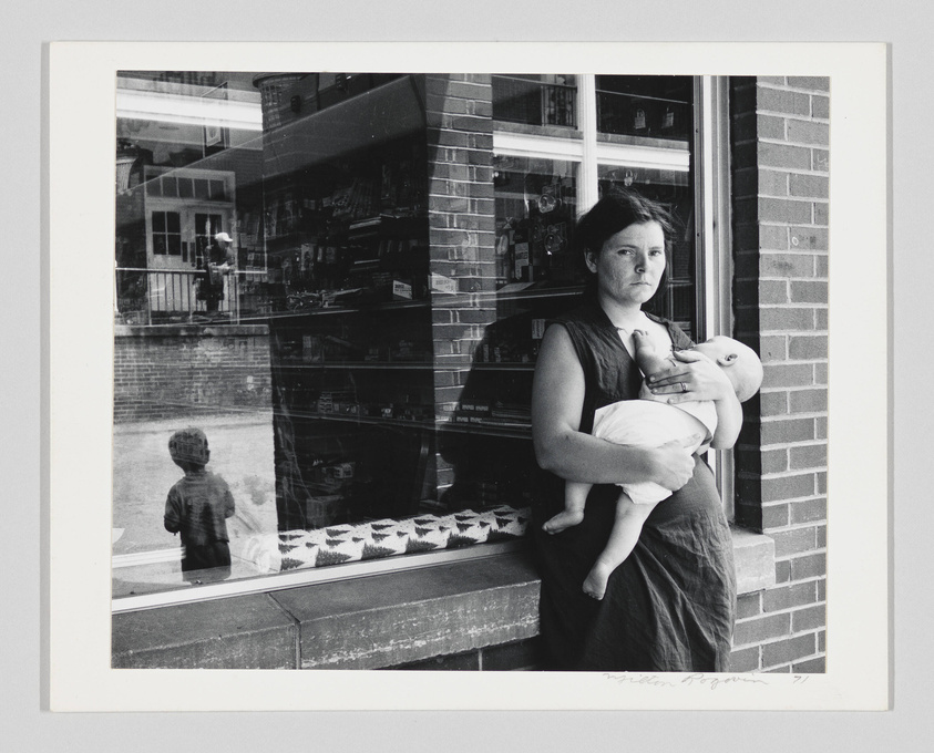 A black and white photograph capturing a woman standing in a doorway holding a baby, with a reflective window to her left showing the image of a child outside and various items displayed inside the shop. The photo has a signature at the bottom right corner.