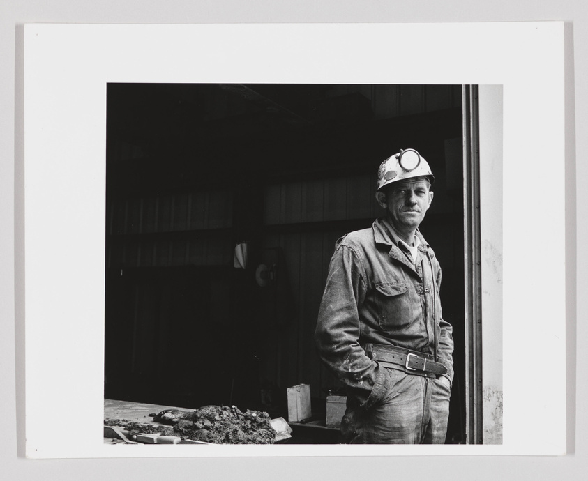 A black and white photograph of a miner standing at the entrance of a building, wearing a hard hat with a headlamp, and a dirtied work uniform. He appears to be taking a break or posing for the photo, with a serious expression on his face.