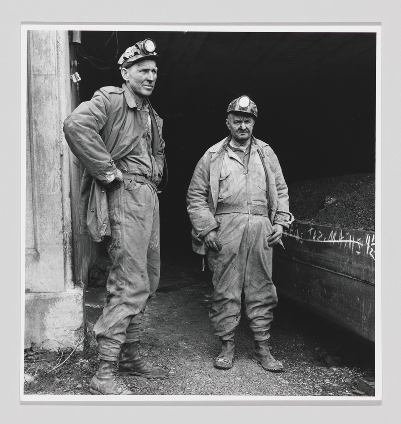 Two coal miners stand at the entrance of a mine shaft. The miner on the left is leaning slightly with his hands on his hips, wearing a headlamp on his helmet, and looking off to the side. The miner on the right stands straight, also with a headlamp on his helmet, looking directly at the camera. Both are wearing work-worn overalls and boots, indicative of a day's work underground.