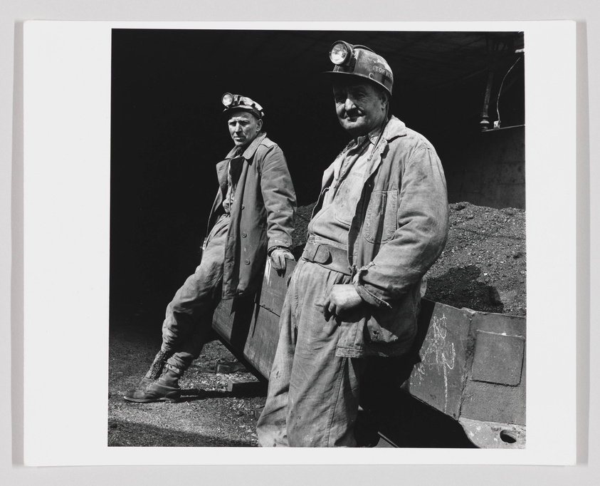 Two coal miners stand at the entrance of a mine, wearing hard hats with lamps and covered in dust, reflecting a moment of rest in their workday.