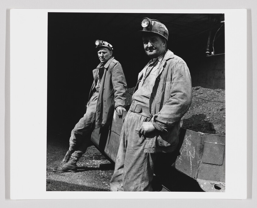 Two coal miners in work clothes and headlamps lean against a coal-filled cart.
