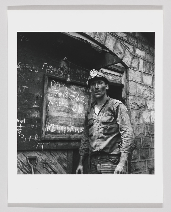 A black and white photograph of a coal miner standing in front of a wooden structure with a chalkboard displaying work-related information. The miner is wearing a hard hat with a headlamp and is covered in coal dust, reflecting a day's work.