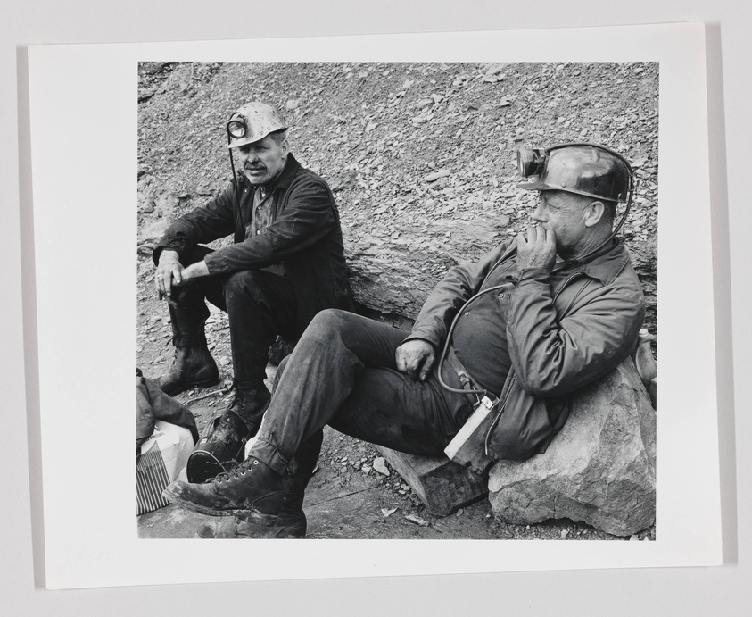 Two miners taking a break, sitting on rocks, with one looking at the camera and the other with his eyes closed, possibly resting. Both are wearing work clothes and helmets with headlamps.