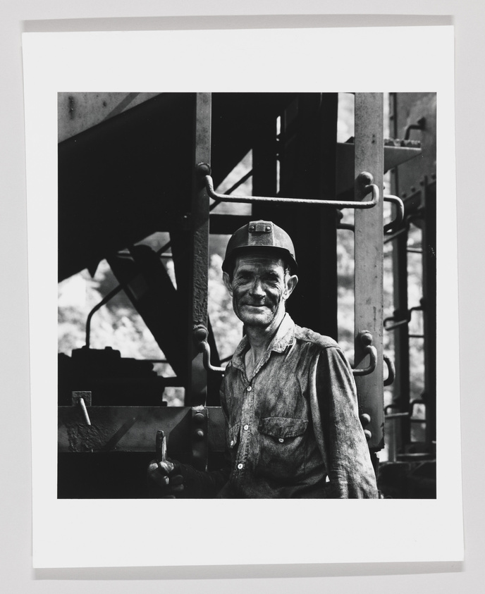 A black and white photograph of a smiling man wearing a hard hat and dirty work clothes, standing in front of industrial machinery. He appears to be a worker taking a break or posing for the photo.