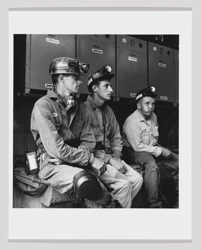 Three coal miners sitting side by side, wearing hard hats with lights and dirty work clothes, appear to be taking a break. They are in front of lockers, looking off to the side, with serious expressions on their faces.