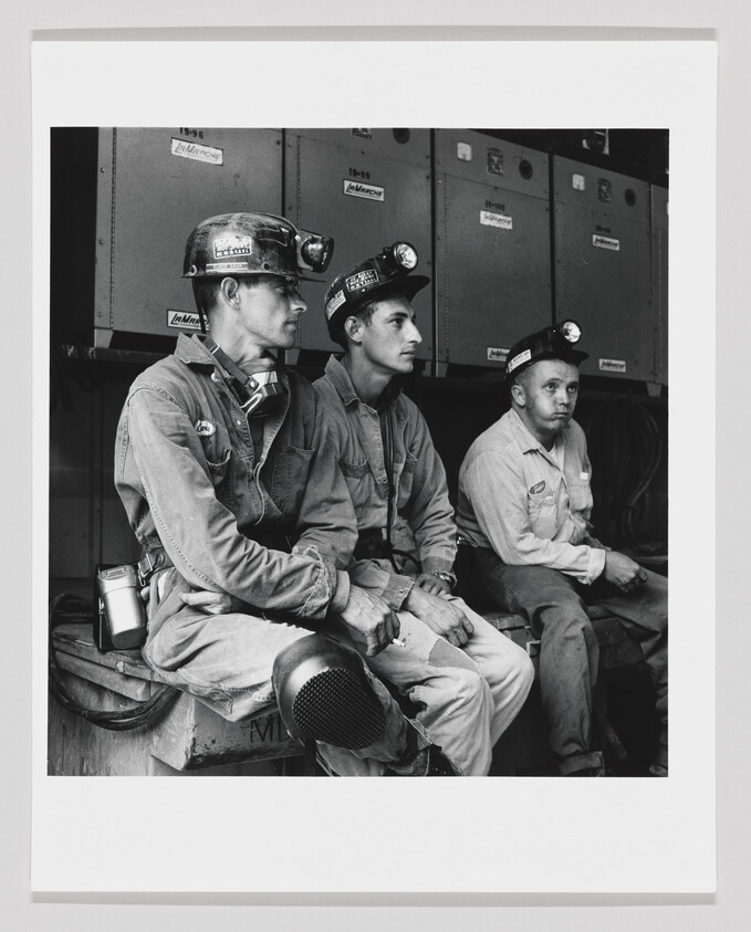 Three miners wearing hard hats with headlamps sit side by side taking a break.