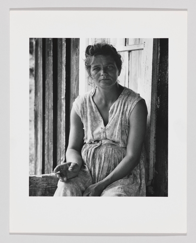 A black and white photograph of a woman sitting in a wooden doorway, wearing a sleeveless dress and looking directly at the camera with a solemn expression. Her hair is pulled back, and she holds a cigarette in her right hand. The image captures a sense of rustic simplicity and quiet strength.