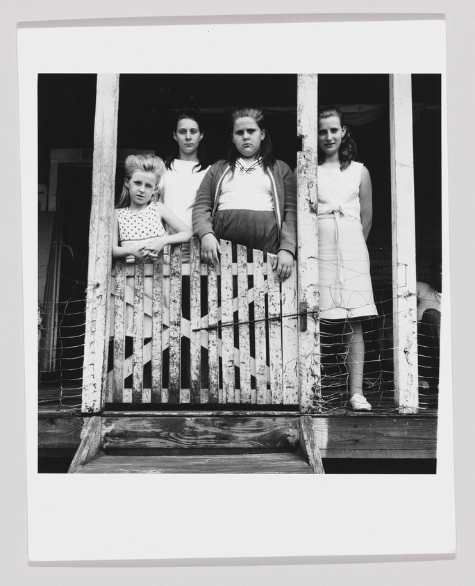 Four young girls standing behind a weathered wooden gate on a porch, with expressions ranging from neutral to slightly serious. The black and white photo captures a moment that feels candid and unposed.