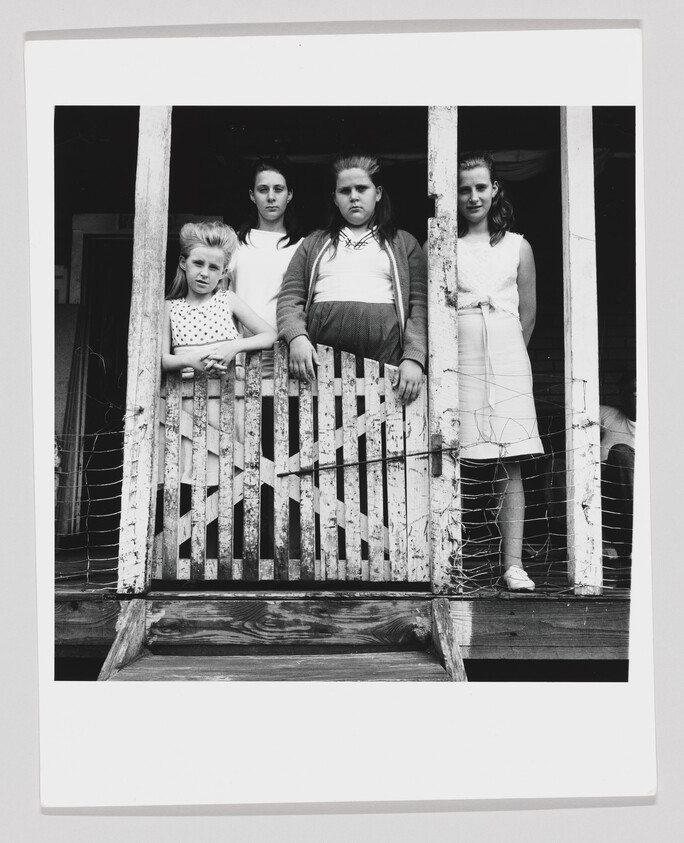 Four girls stand behind a worn wooden gate on a porch, looking directly at the camera.