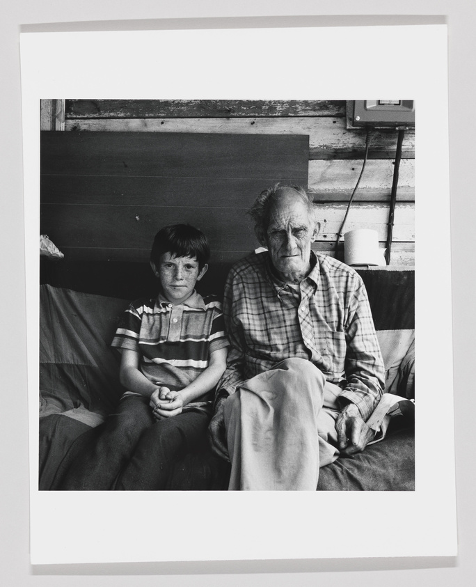 A black and white photograph of a young boy and an elderly man sitting side by side on a couch, with the boy to the left looking at the camera and the man to the right looking down slightly. Both are dressed casually, the boy in a striped polo shirt and the man in a plaid shirt. The background features a wooden wall and a thermostat, with a roll of paper towels resting on a ledge.