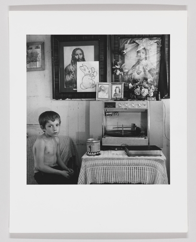 A black and white photograph featuring a young boy sitting shirtless on a chair to the left, with a contemplative expression. Behind him, a wall adorned with religious imagery, including a framed picture of Jesus and a veiled woman, as well as a child's drawing of a rabbit. To the right, an old-fashioned record player sits atop a lace-covered table, with a can of Bugler tobacco and a small radio beside it. The setting suggests a modest, personal living space.