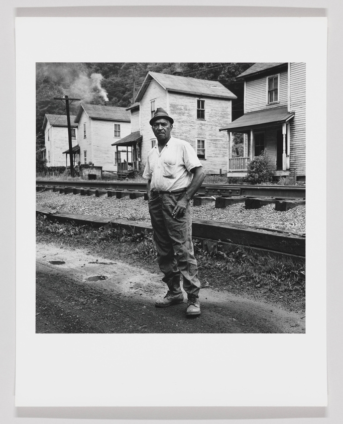 A black and white photograph of a man standing confidently with his hands on his hips next to railroad tracks. He is wearing a cap, a short-sleeved shirt, and work pants. Behind him are two-story houses and a smokestack emitting smoke, suggesting an industrial or rural setting.